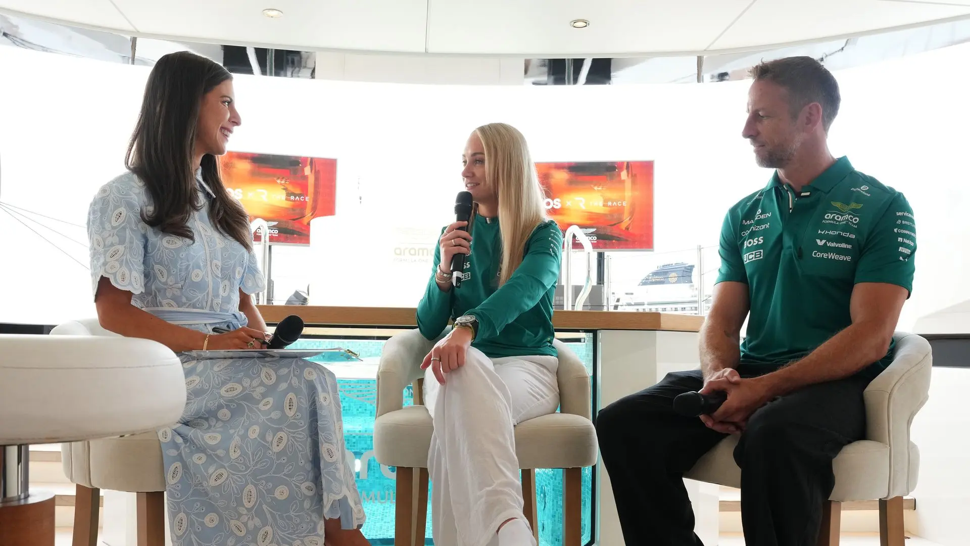 Three people sit on stools in a bright, modern yacht interior; left woman in a blue lace dress holds a microphone, middle blonde in teal speaks, right man in teal polo listens.