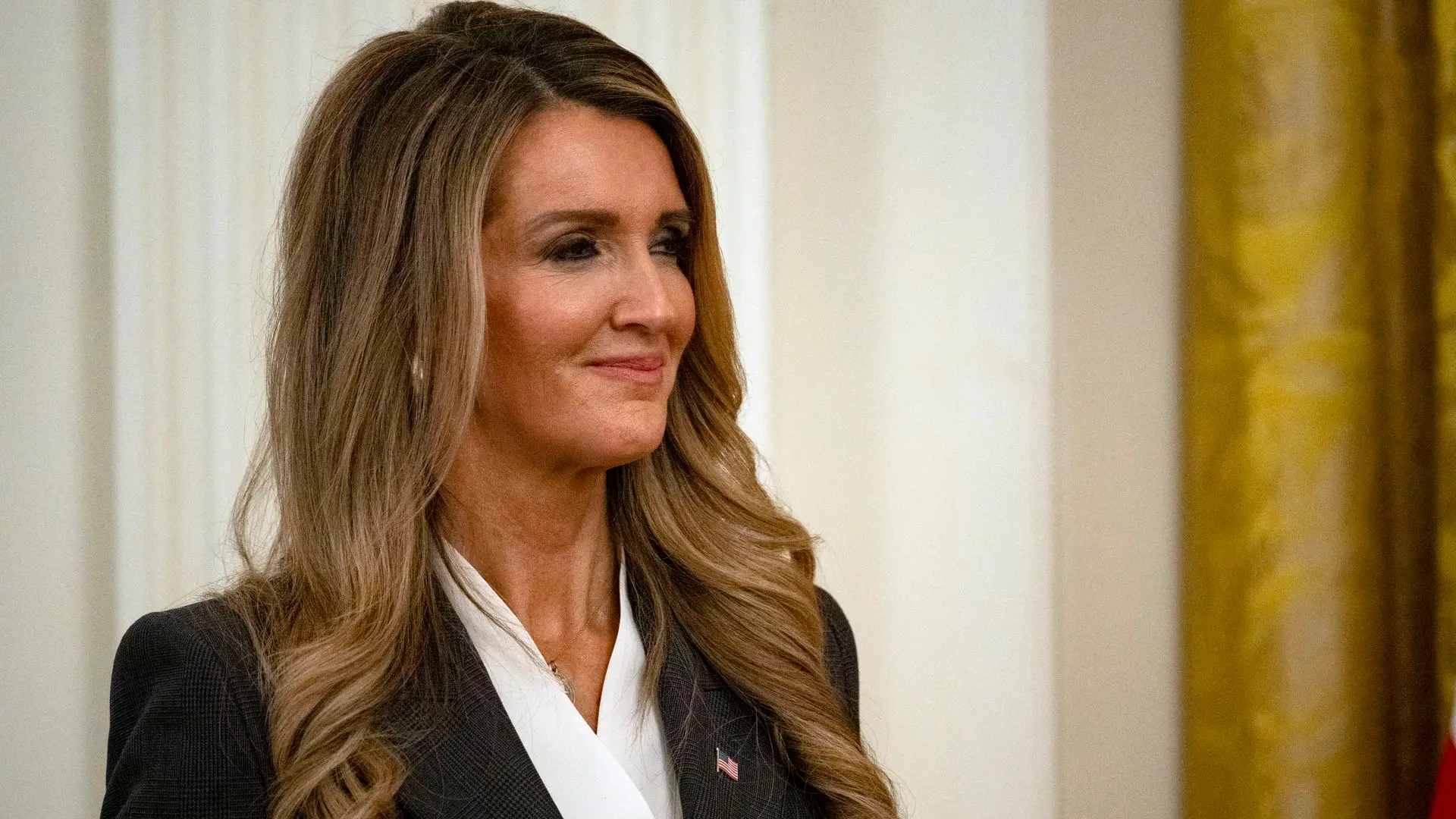 Kelly Loeffler smiles as she looks on while inside the White House during an event. She is wearing a dark blazer, a white blouse and an American flag pin, and her hair is down.