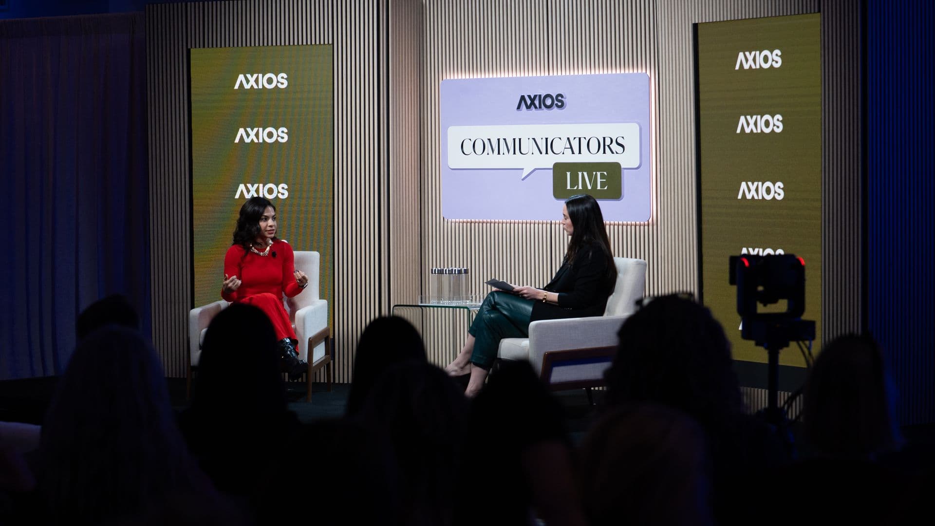 Two women seated on a stage having a discussion at an Axios event called "Communicators Live," with an audience in the foreground and Axios logos on yellow vertical screens.