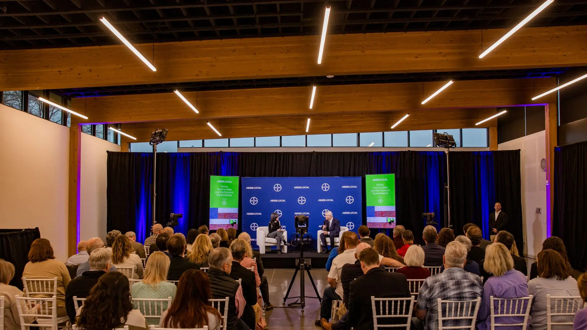 Panel discussion in a modern hall with wooden beams. Two speakers on stage by a blue backdrop with green banners, facing a seated audience of white chairs.