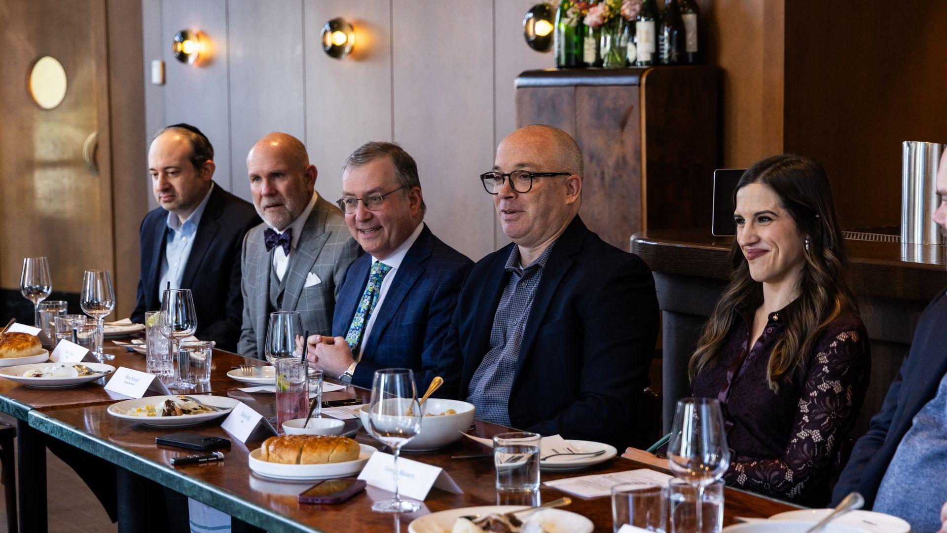Five adults seated at a dining table with plates, glasses, and bread in a warmly lit room with wood paneling, engaged in conversation and smiling.