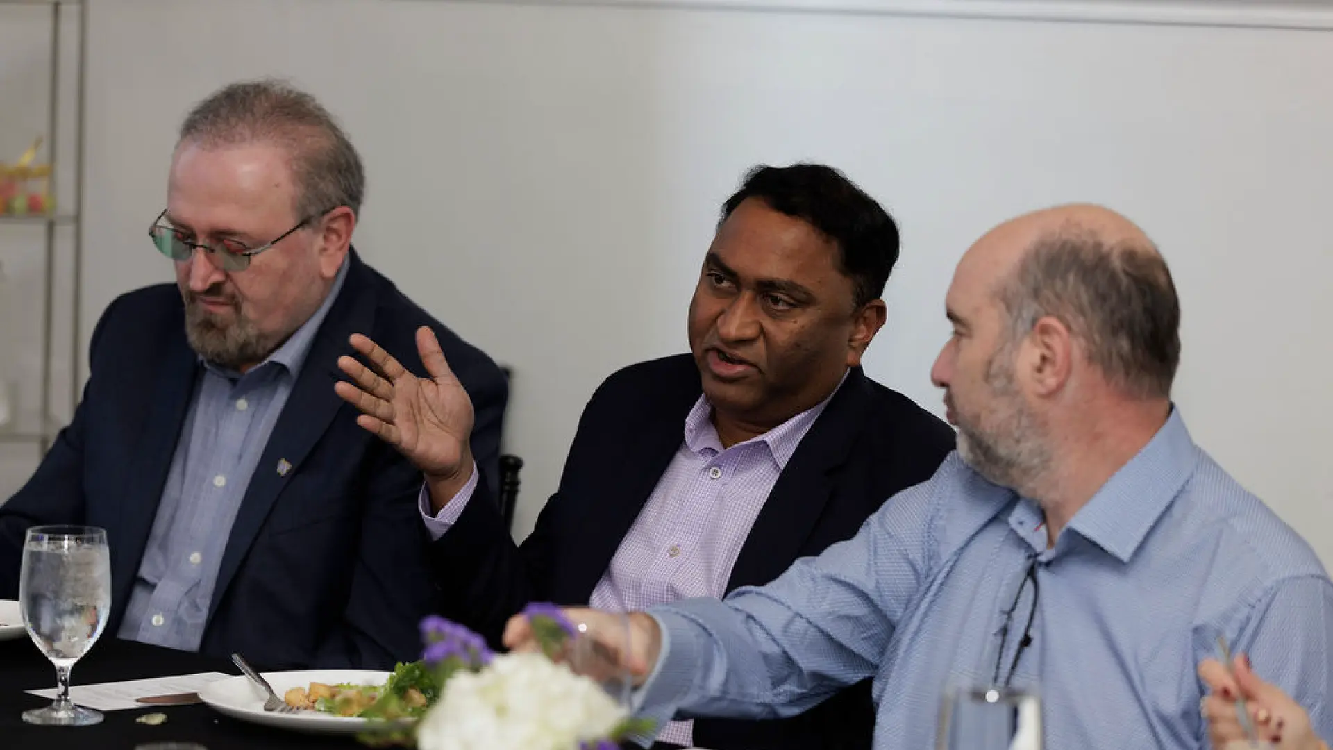 Three men in business attire sitting at a table with plates of food and glasses of water, engaged in conversation in a meeting or dining setting with a flower centerpiece.