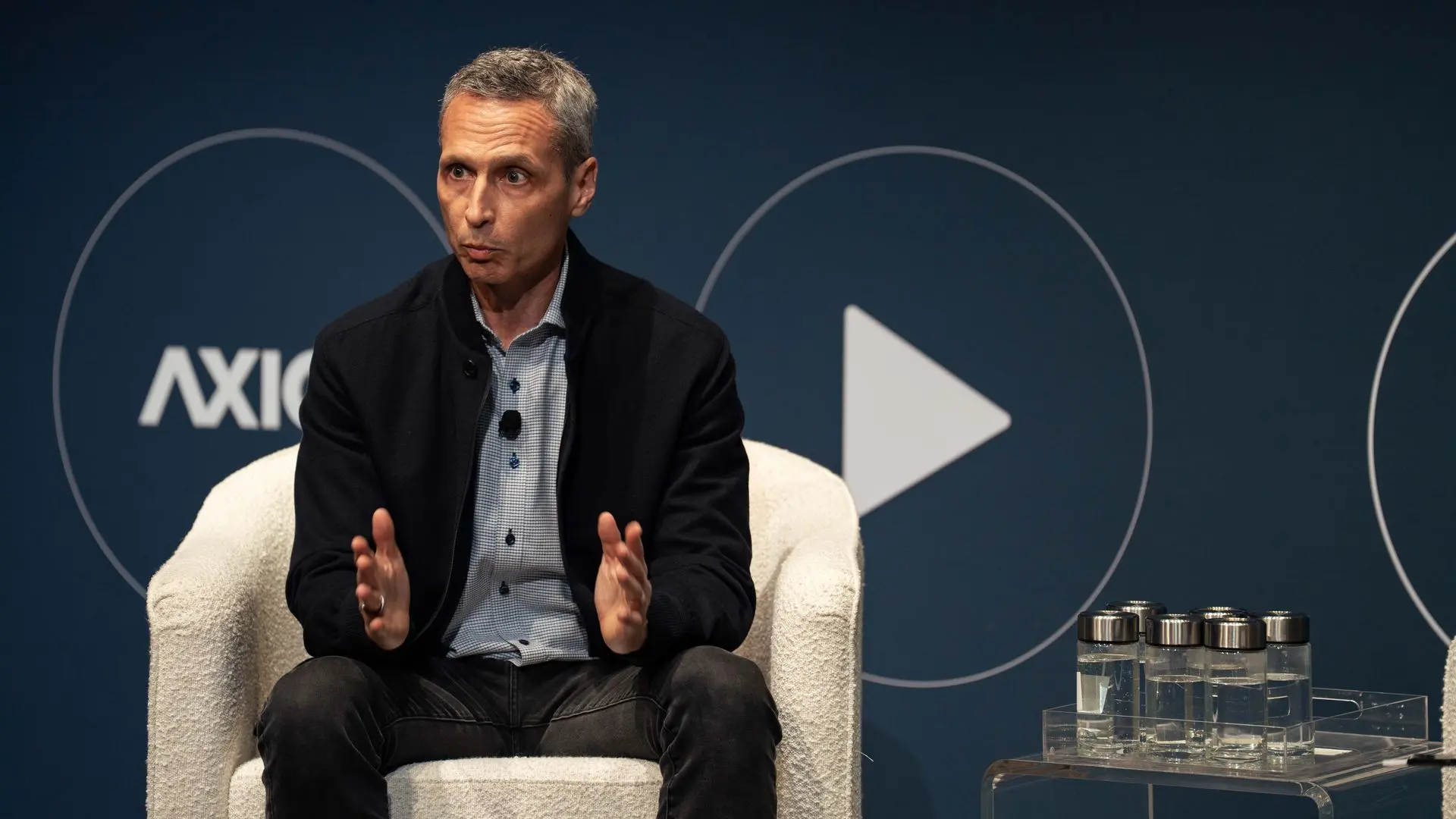 ESPN chair Jimmy Pitaro in a black jacket and checkered shirt speaking while sitting in a white chair on stage, dark blue background at Media Trends Live.