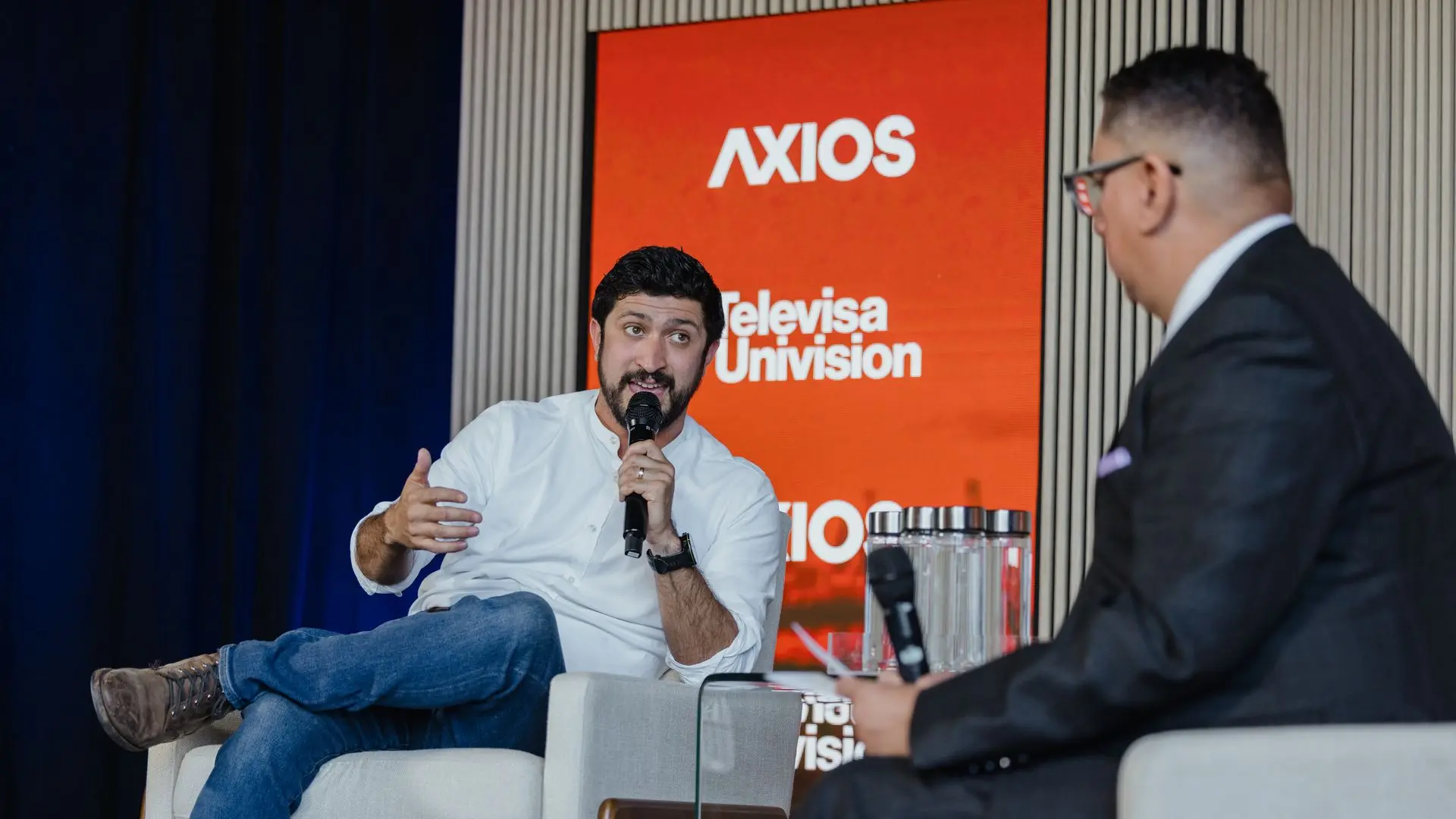Two men interview on stage; left man in white shirt and jeans speaks into a microphone while gesturing, right man in a suit listens. An orange backdrop displays AXIOS and Televisa Univision logos.