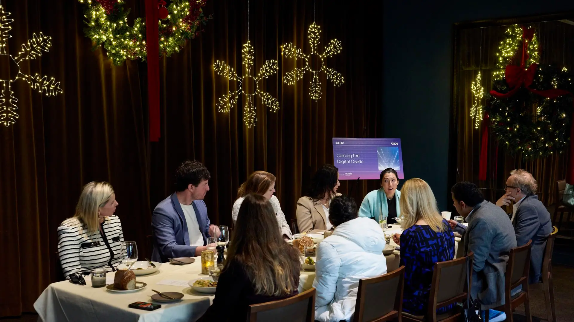 Group of nine people seated around a table with food, discussing in a warmly lit room decorated with large lighted snowflakes and Christmas wreaths with red bows. A sign reads "Closing the Digital Divide."