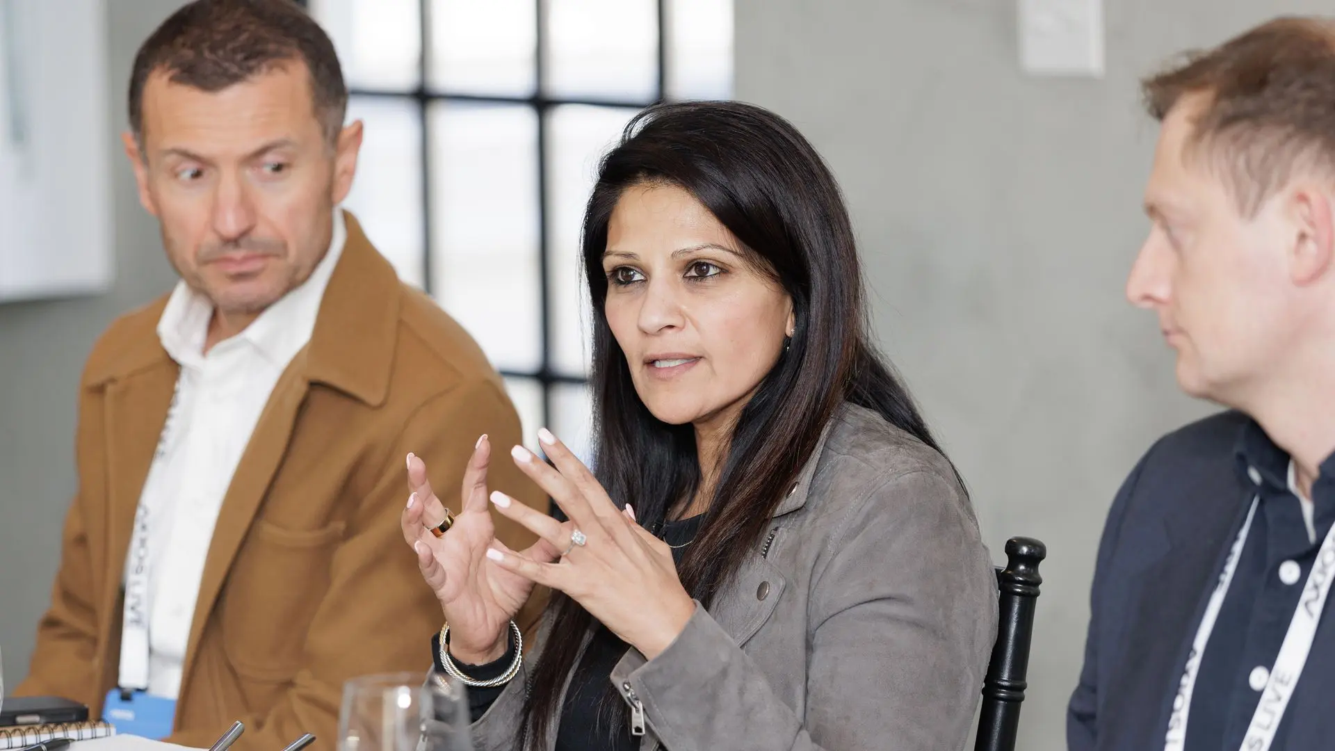 Three people sit at a table having a discussion. The woman in the center, with dark hair and a gray jacket, speaks using hand gestures. Two men, one in a brown jacket and one in navy, listen attentively.