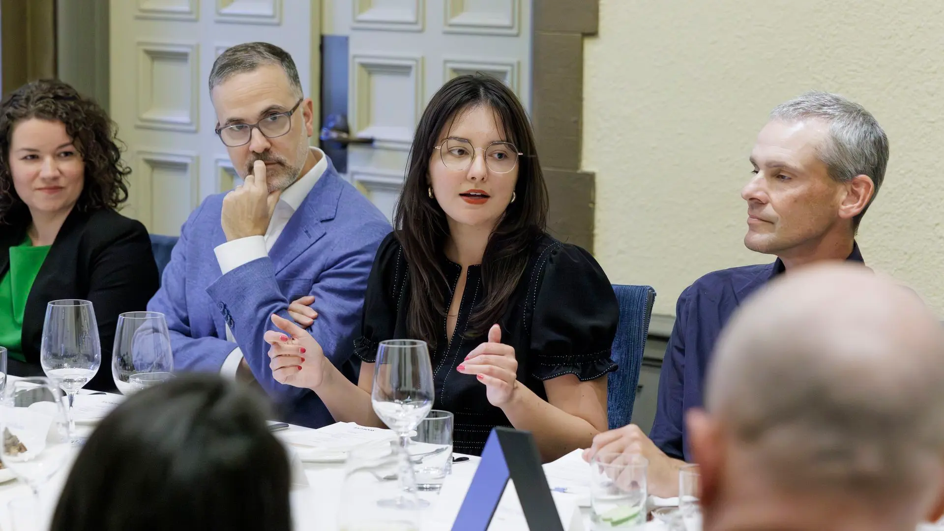 Four people sit at a formal round-table; the center woman with glasses in a black blouse speaks, flanked by a man in a blue blazer and another man, with a woman in green on the left.