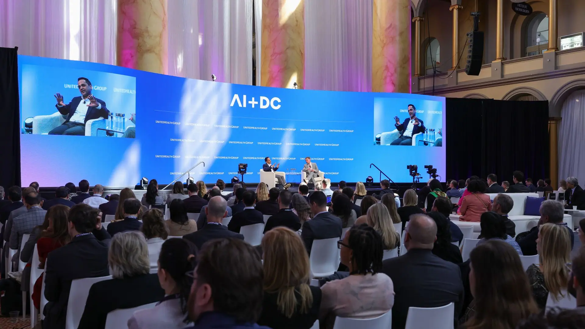 Large conference hall with blue stage backdrop reading "AI+DC". Two people seated on stage in conversation, audience facing them. Side screens show the speaker. Marble columns, purple lighting.