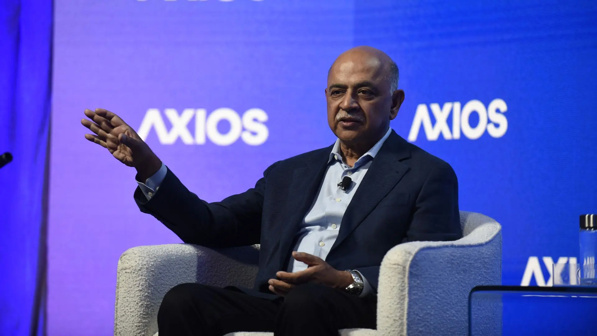 Bald man in a dark suit and light shirt sits in a white armchair on a blue stage, gesturing with his left hand; a blue backdrop behind shows the word "AXIOS".