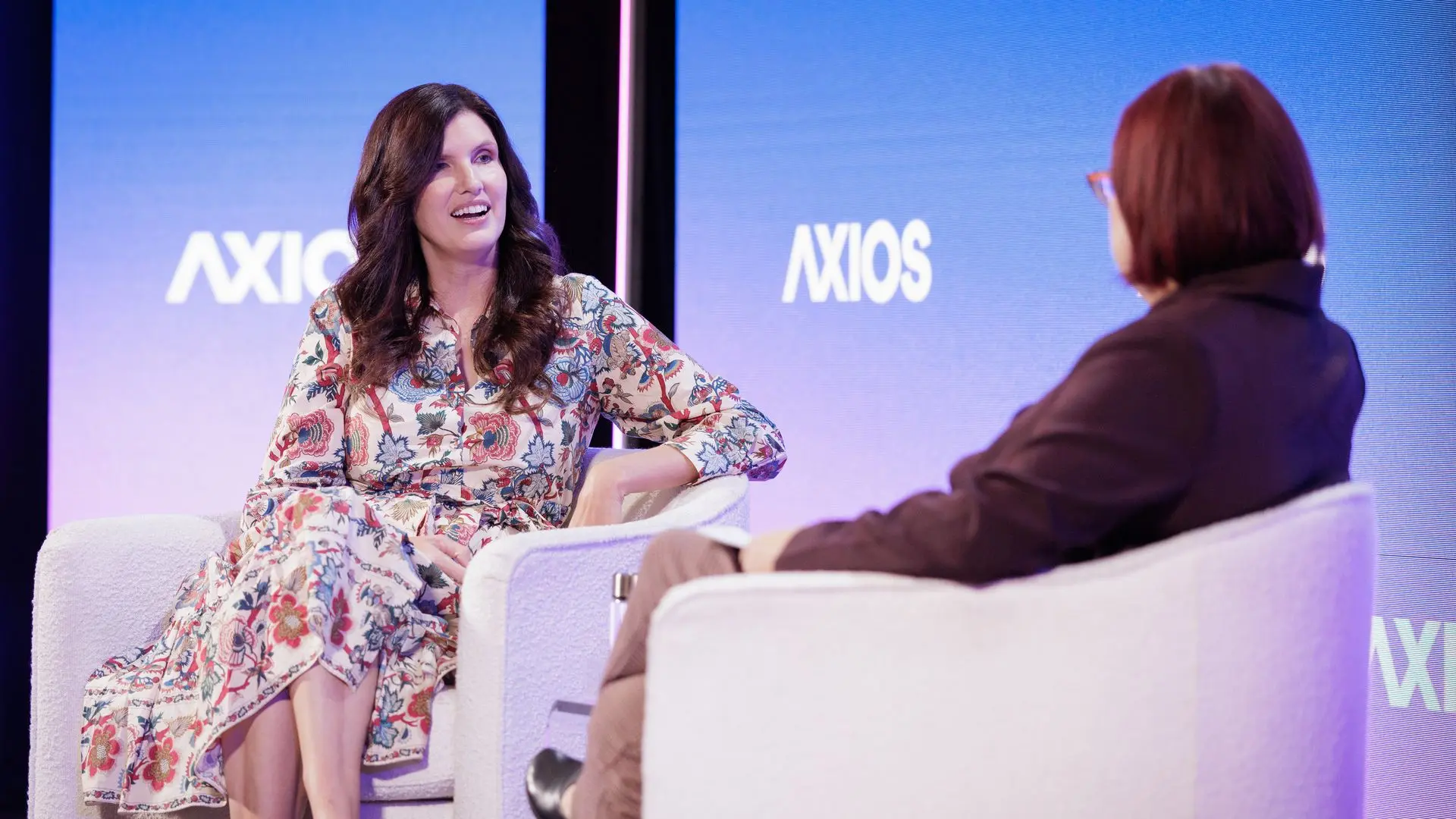 Two women seated in white chairs on stage at an Axios event, one woman with dark hair wears a floral dress, the other with reddish hair wears a brown outfit, blue Axios logos in background.
