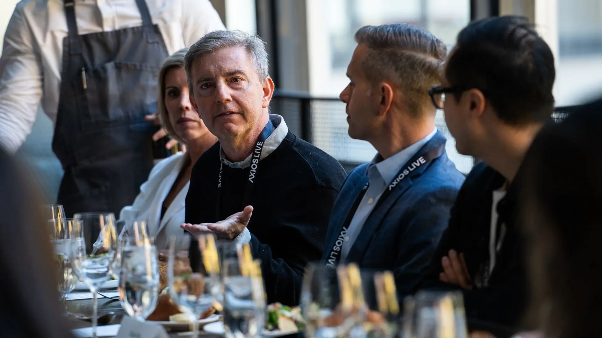Group of professionally dressed people wearing "AXIOS LIVE" lanyards seated at a table with water glasses and plates. One man is speaking with a hand gesture.