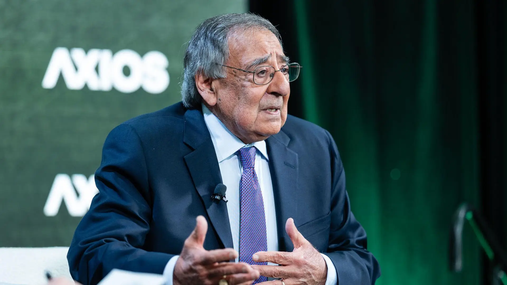 Man in dark suit and purple tie speaks during an Axios event, gesturing with hands, green backdrop and Axios logo blurred behind him.