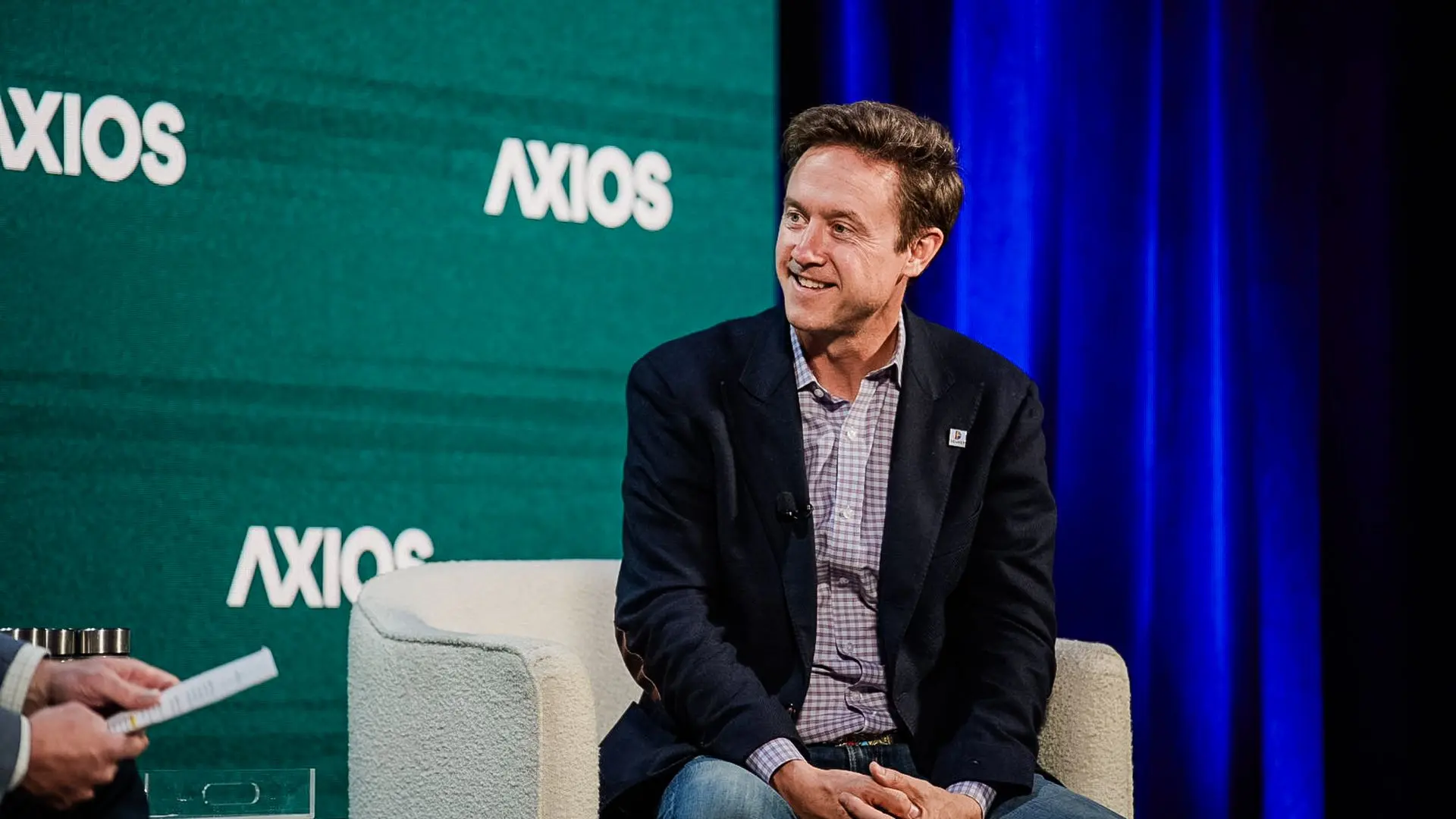 A man in a dark blazer and checkered shirt sits on a light gray chair on stage, smiling, with a green Axios backdrop and blue curtains to the side.
