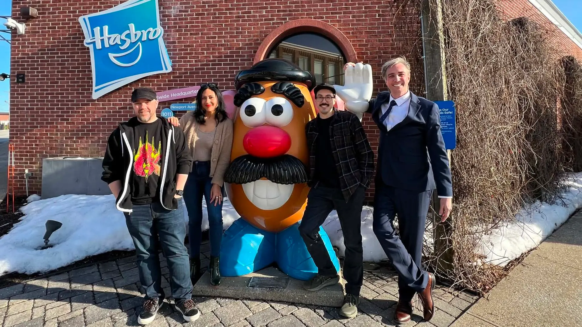 Five people stand in front of a red brick Hasbro building with a large Mr. Potato Head statue. Clear blue sky, patches of snow, and bare vines along the wall.