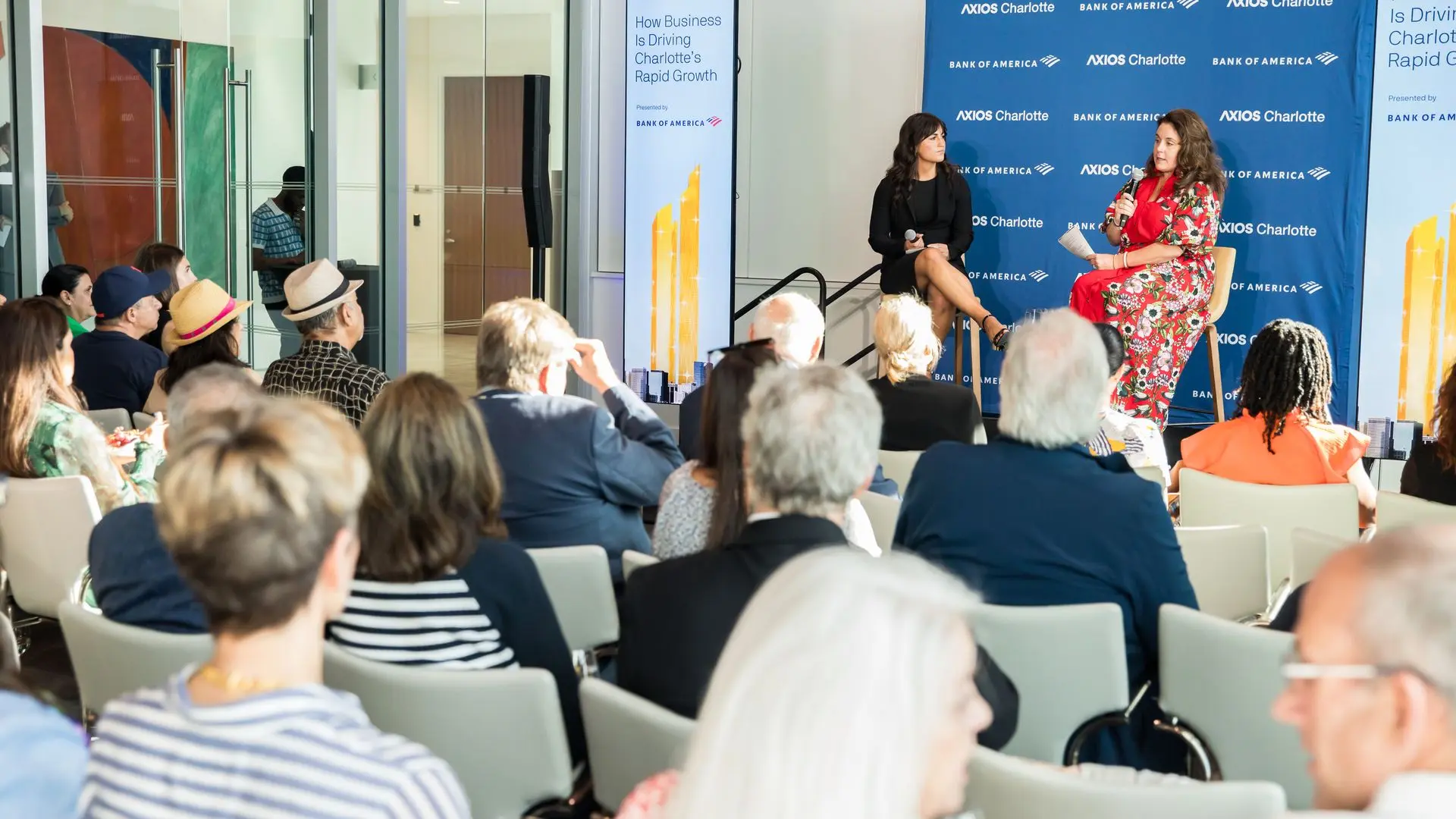Panel discussion with two women on stage: one in black, the other in red floral dress with a microphone, before a blue Axios Charlotte backdrop and seated audience.