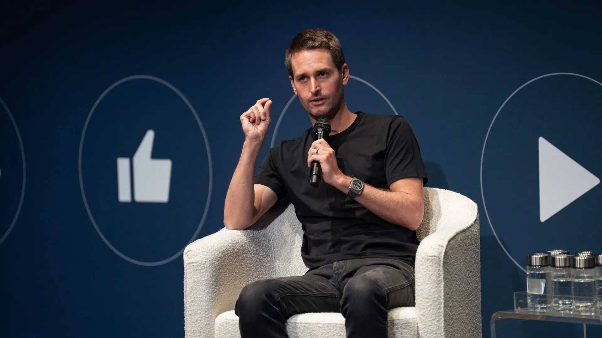 Snap CEO Evan Spiegel in a black t-shirt and jeans holding a microphone, sitting on a white armchair against a dark blue background with large white thumbs-up and play icons.
