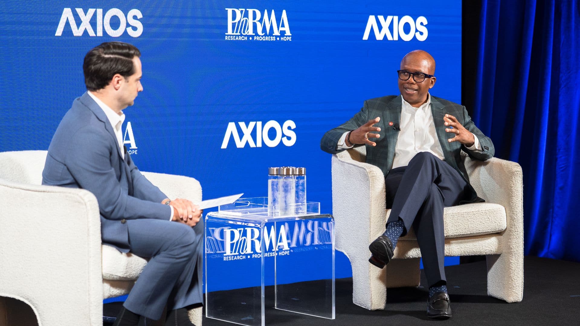 Two men in suits sit on beige chairs on a stage with a blue Axios backdrop; one gestures as they talk, a glass table and water pitcher between them.