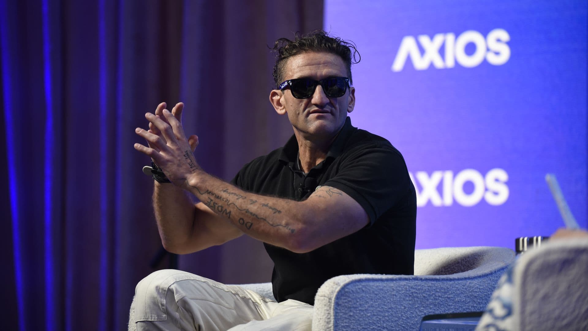 Man wearing a black shirt and sunglasses sits on a blue stage with a purple curtain on the left, gesturing with his hands. A blue backdrop shows the word AXIOS in white.