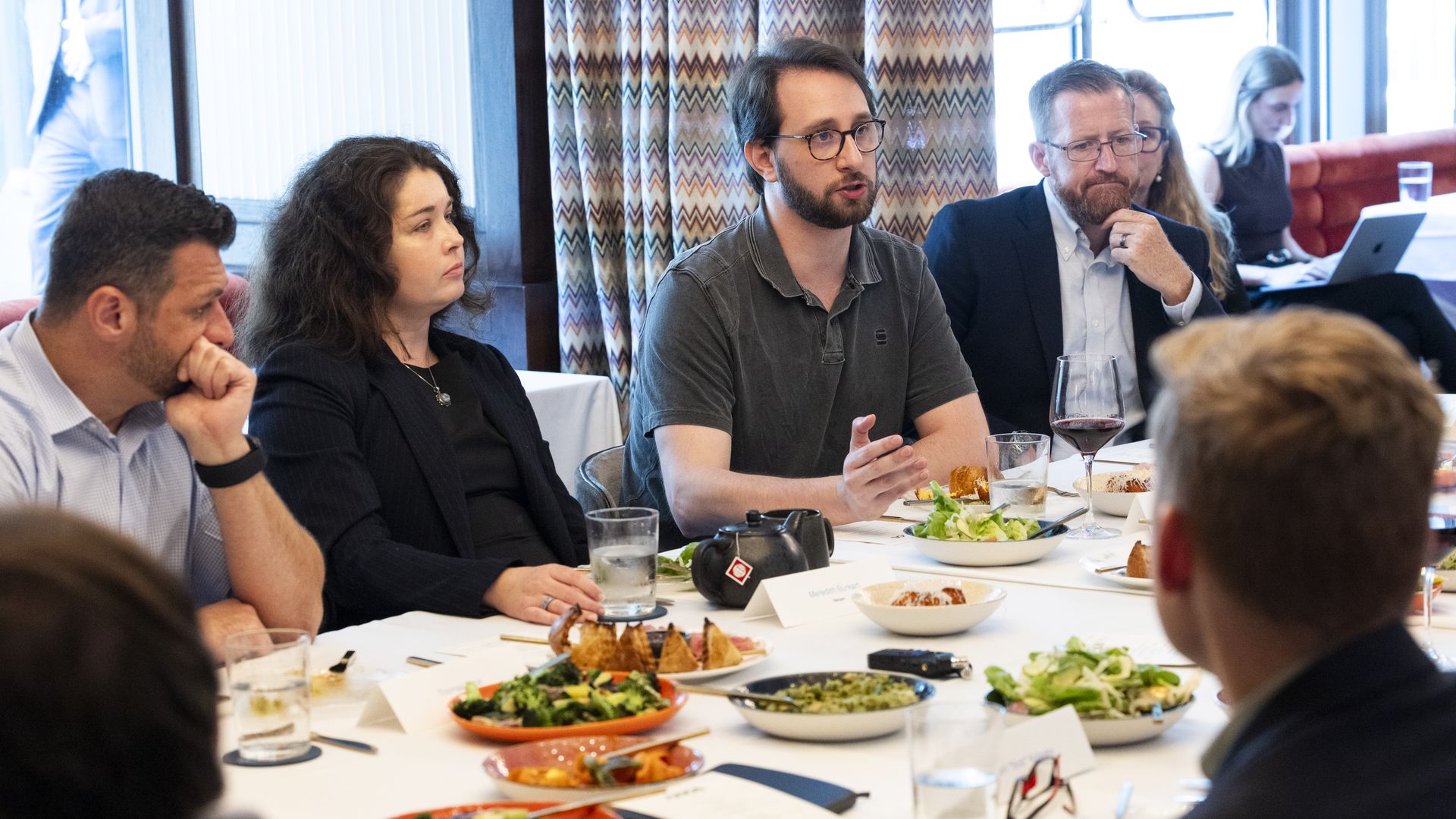 Professionals at a long dining table in a bright restaurant. A man in a gray polo speaks as others listen; dishes include salad and bread, with wine glasses and a laptop visible.