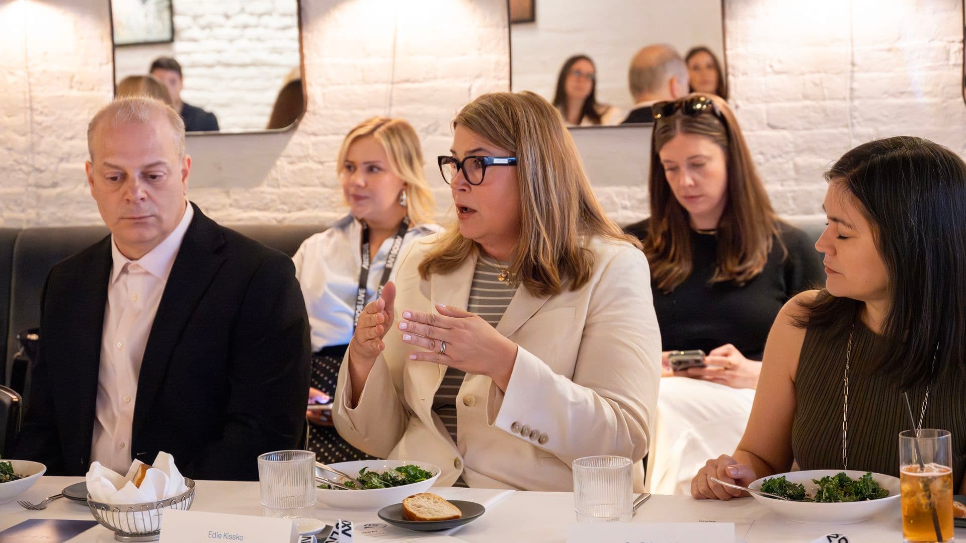 Business meeting in a bright restaurant: a woman with glasses speaks animatedly at a table with salads, bread, and water, others listening; white brick walls and modern mirrors in the background.