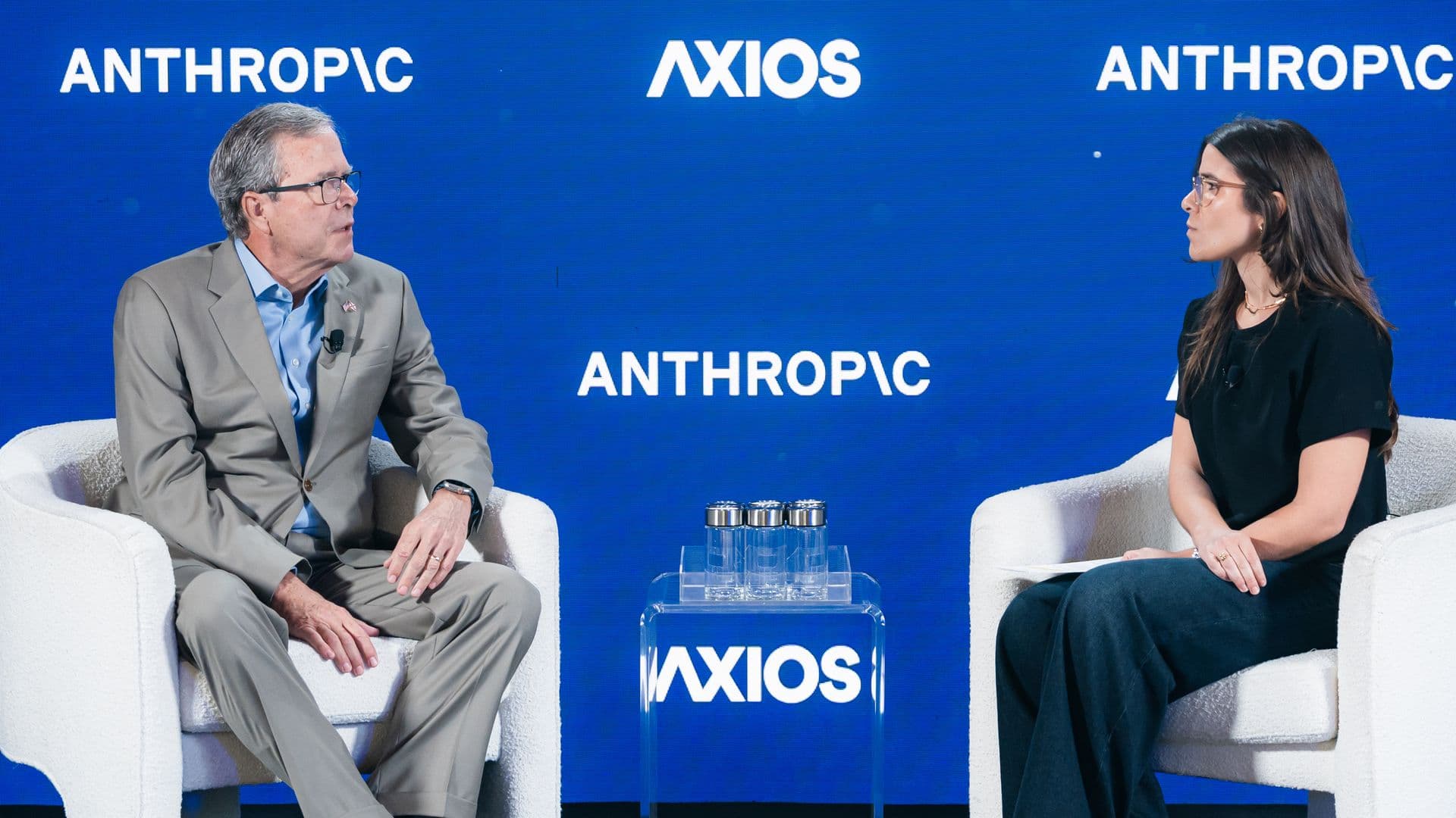 Panel discussion on a blue backdrop with AXIOS and ANTHROPIC logos; a man in a gray suit and a woman with dark hair and glasses sit on white chairs, microphones clipped, small glass table between them.