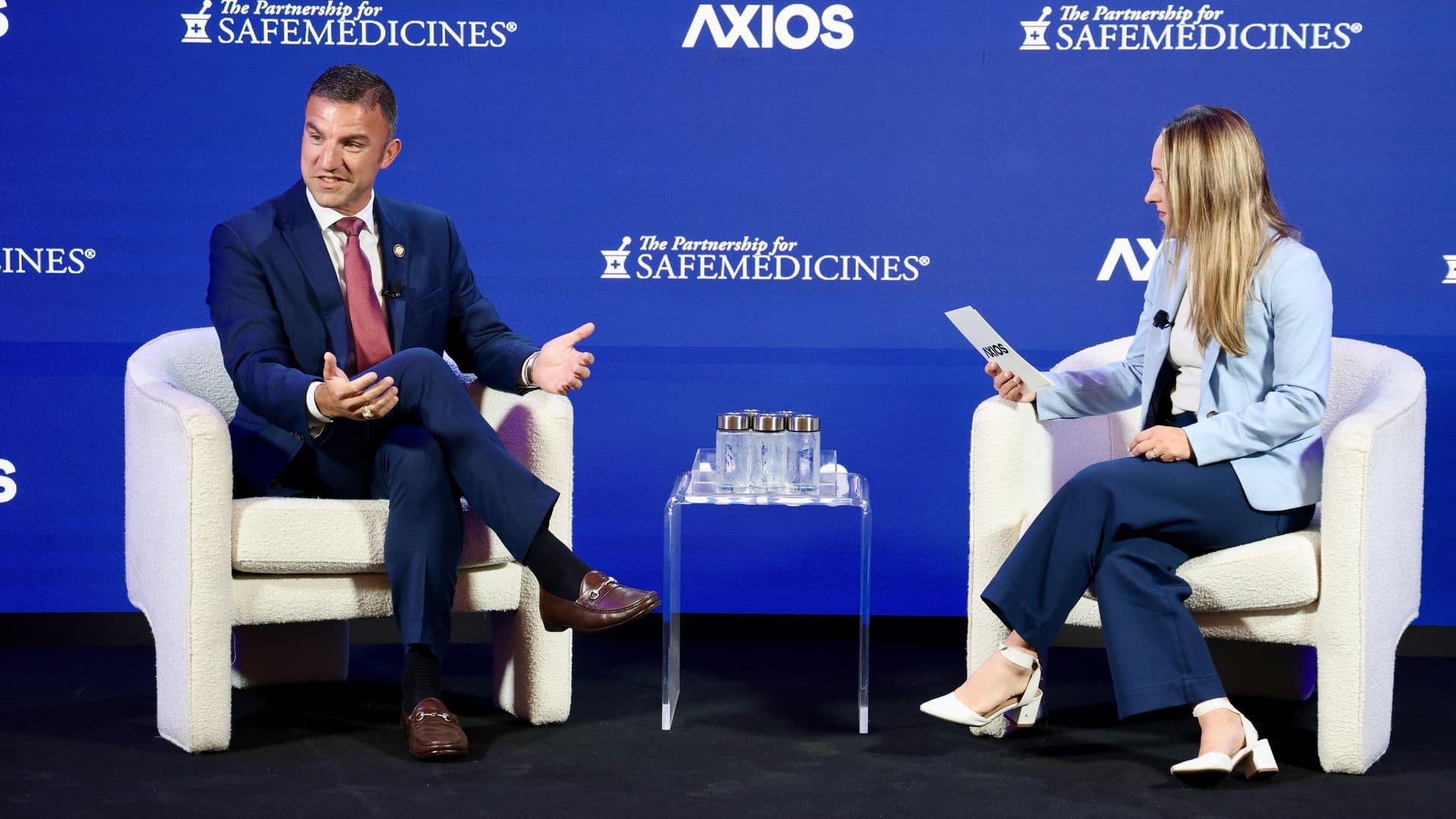 Two panelists on a stage backdrop with Axios and Safemedicines logos. A man in a navy suit gestures while speaking; a woman in a light-blue blazer holds a card beside a glass table.