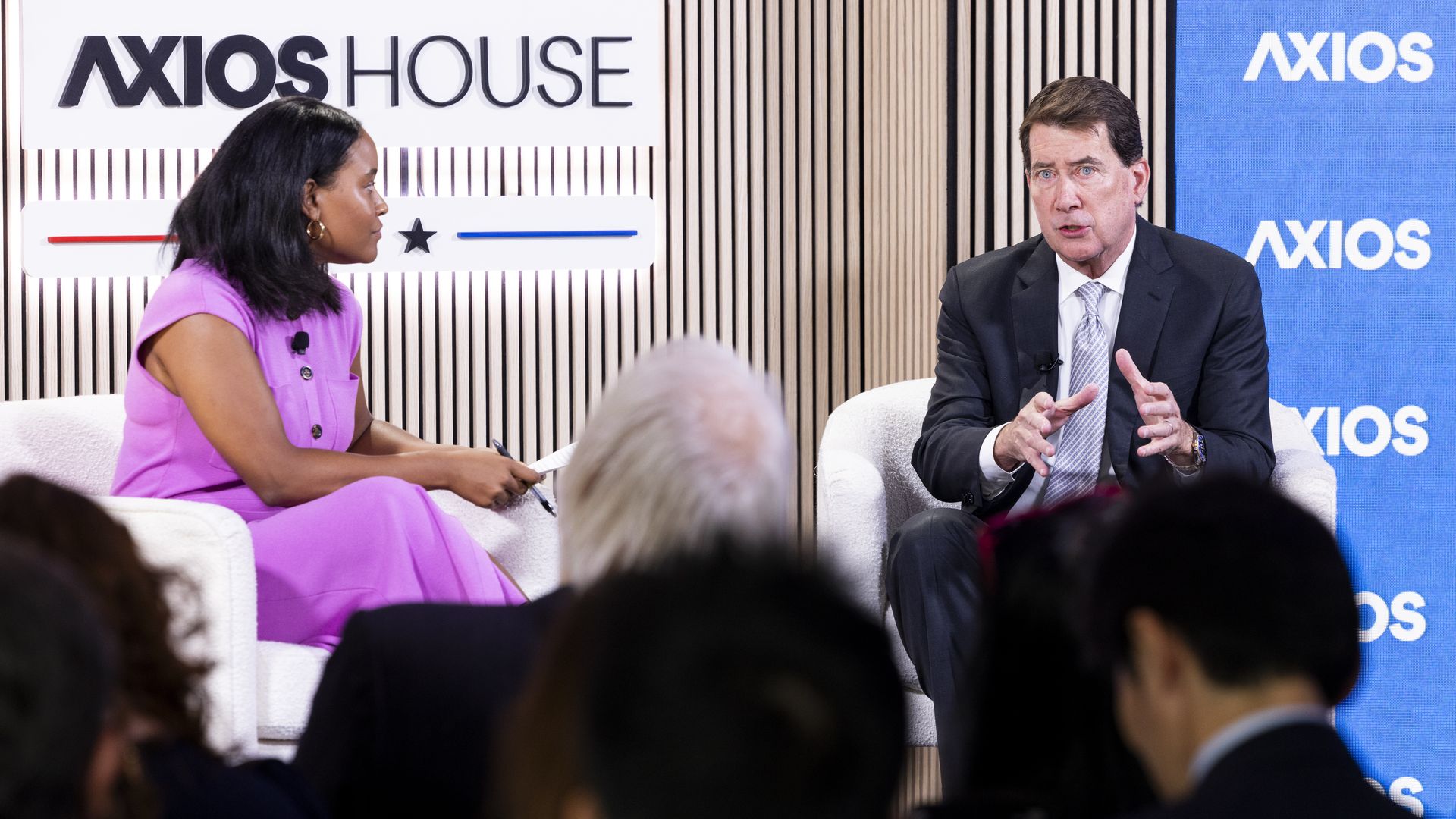 Two people sit on white chairs in a studio: a woman in a bright pink dress on the left and a man in a dark suit on the right, with "AXIOS HOUSE" signage and a blue Axios backdrop.