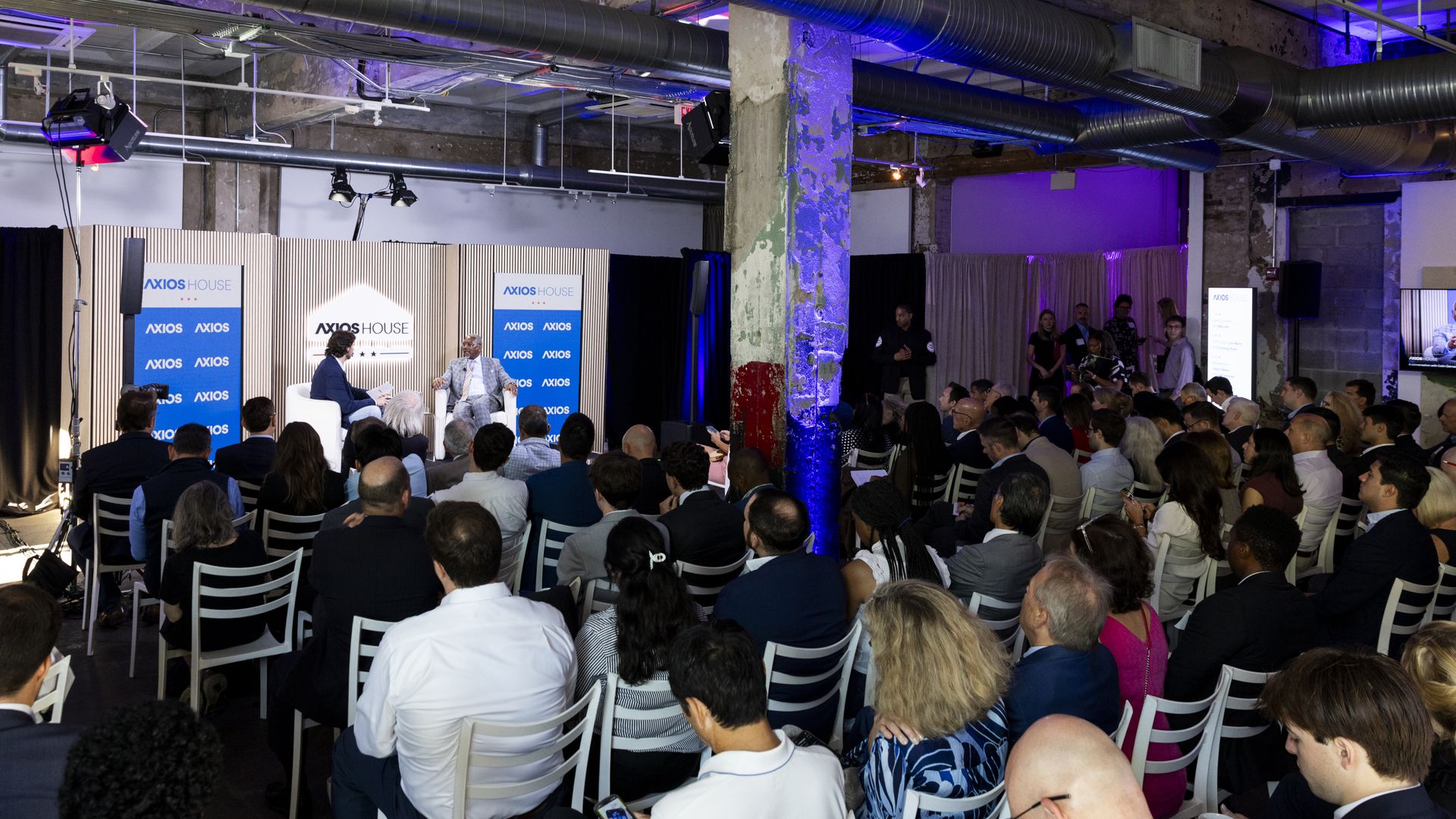 An indoor panel discussion in a warehouse-like venue. Two speakers sit on a stage with "Axios" branding behind them, while a large audience watches, surrounded by exposed ducts and purple lighting.