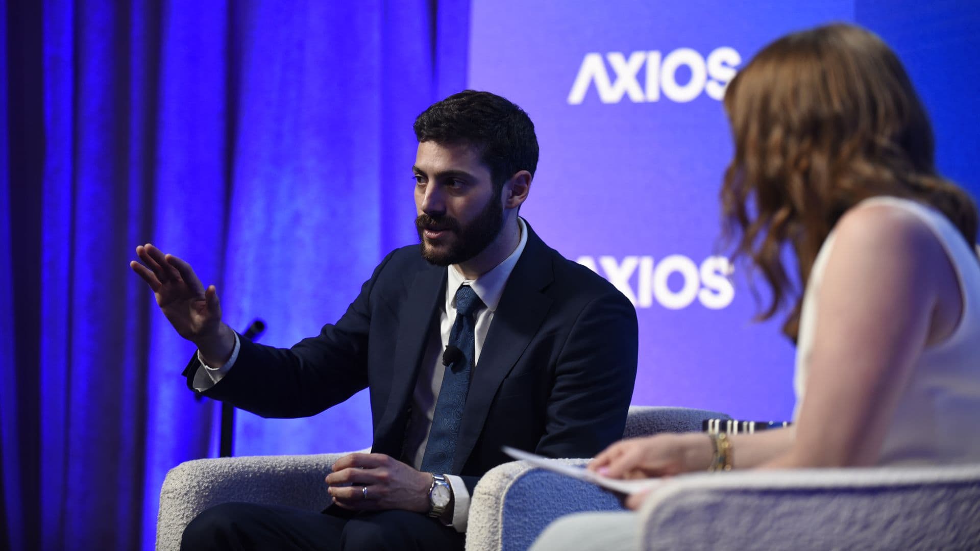 Bearded man in a dark suit speaks and gestures on a blue-lit stage; a woman with light hair sits nearby. A large Axios logo appears on the blue backdrop.