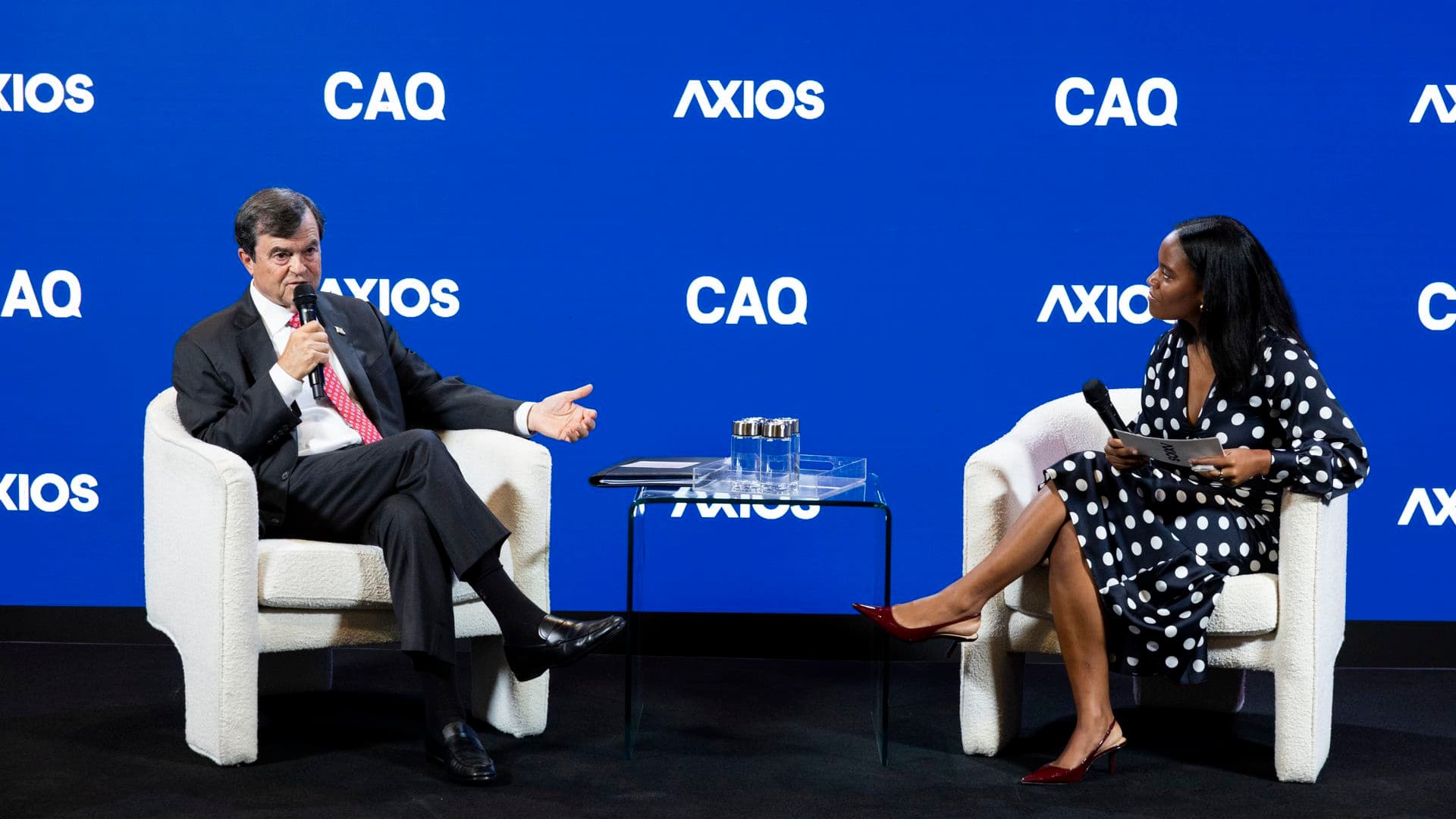 Two people sit in white armchairs on stage with backdrop reading "AXIOS" and "CAQ." A man speaks into a microphone while a woman in polka-dot dress listens; a glass table sits between them.