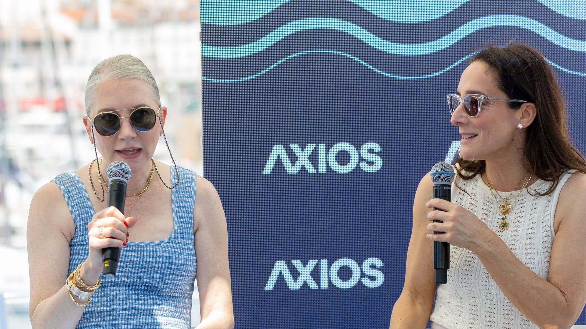 Two women in sunglasses hold microphones at an outdoor event, speaking in front of a blue digital backdrop showing waves and the word AXIOS. Left, silver hair in a blue check dress; right, white top.