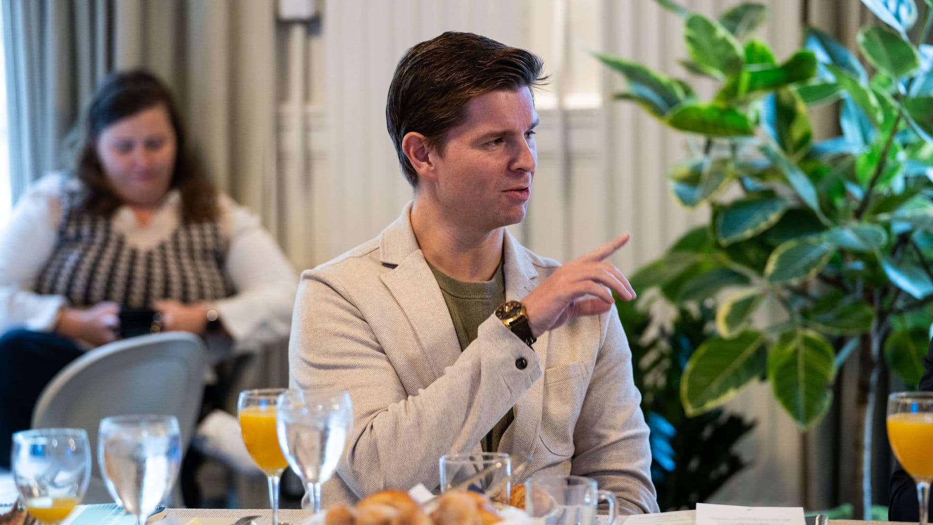 Man in a beige blazer sits at a table, gesturing with his hand. A blurred woman sits in the background; large green plants on the right. Tableware includes glasses, orange juice, and bread.