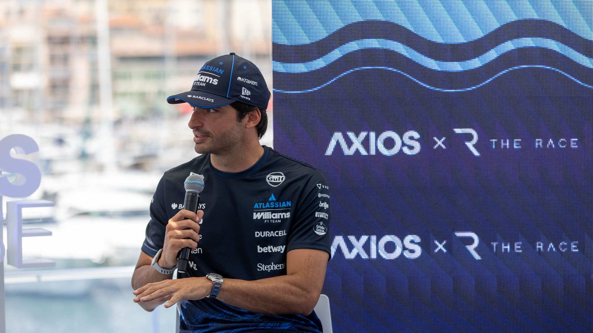 Carlos Sainz wearing a dark blue cap and shirt holds a microphone beside a large blue Axios display with wave patterns, sponsor logos on his clothing visible.