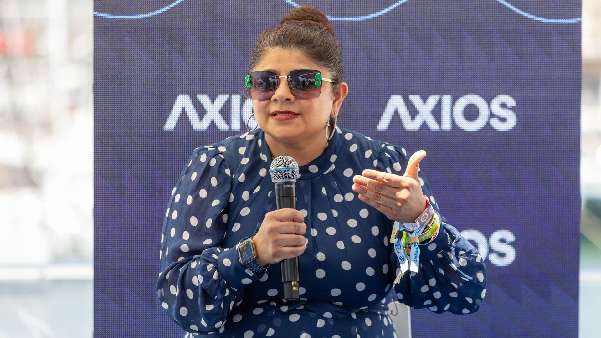 Woman with dark hair in a top bun, wearing oversized sunglasses with green accents, a navy polka-dot dress, hoop earrings, and a smartwatch, holding a microphone in front of an Axios backdrop.