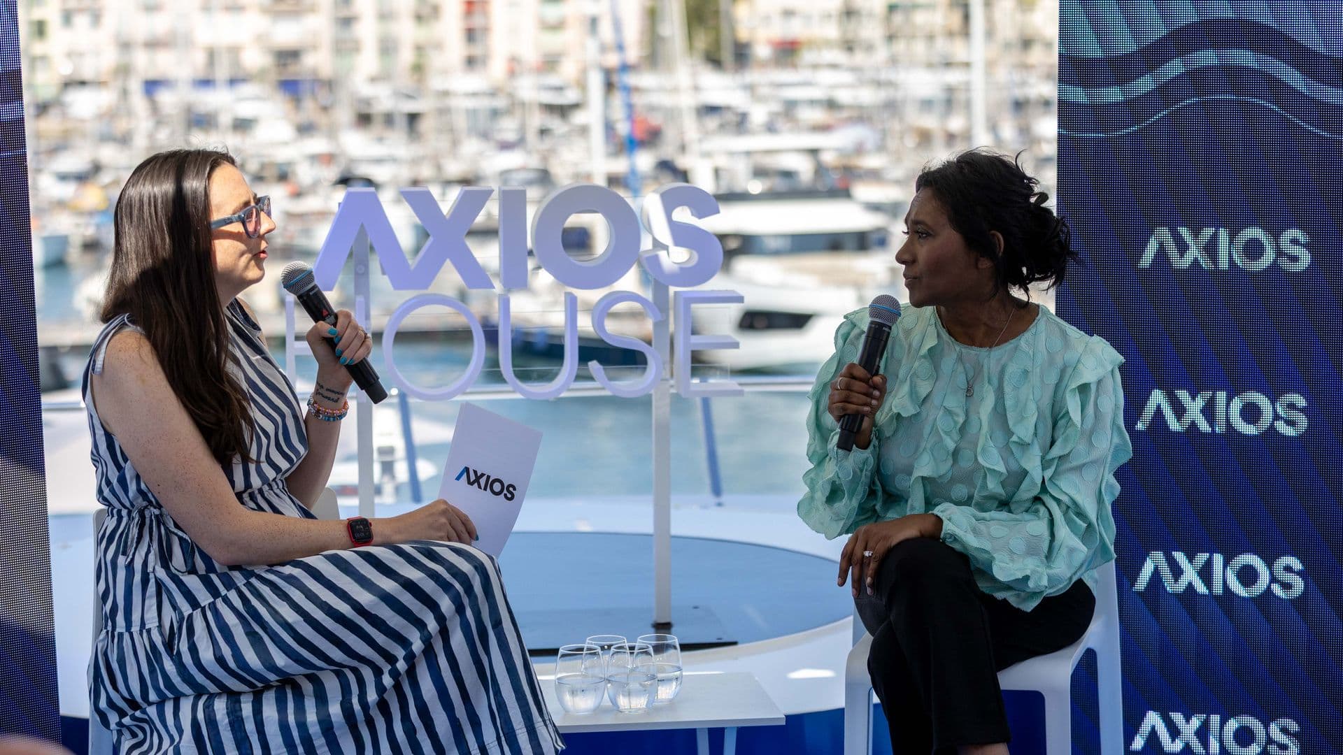 Two women on a panel by a marina, each holding a microphone. Left wears a striped blue-and-white dress and glasses; right wears a mint green blouse. Large AXIOS signs and boats in background.